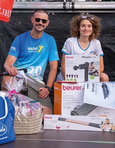 Two adults wearing casual sportswear sit next to each other with various boxed fitness and wellness products and a gift basket in front of them. Beschreibung von KI generiert