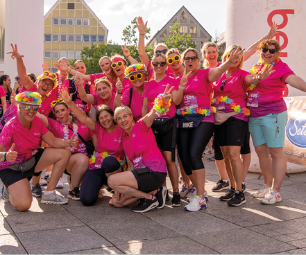 A group of people wearing matching bright pink shirts and colorful accessories are posing energetically outdoors in front of a large inflatable arch and buildings. Beschreibung von KI generiert