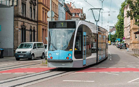A modern blue and white tram is traveling on tracks through a city street lined with buildings and parked vehicles. Beschreibung von KI generiert