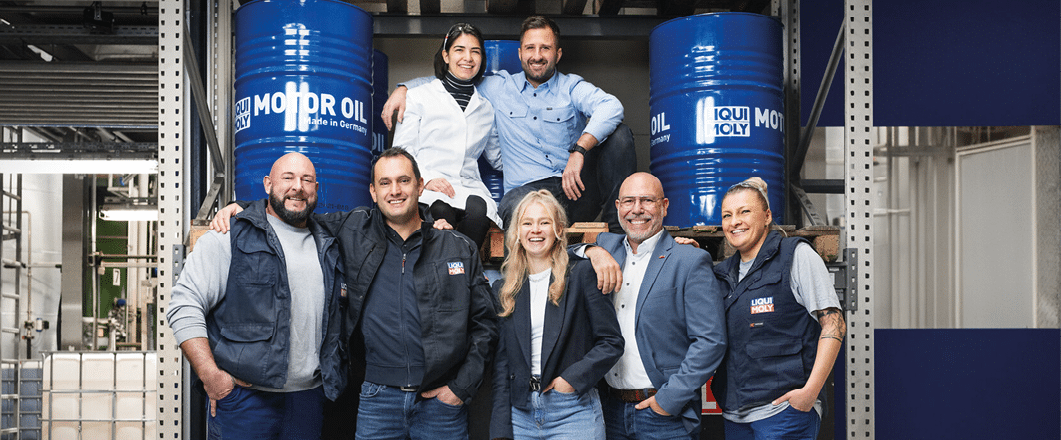 Five people wearing casual and work clothing stand side by side in an industrial warehouse setting with large containers in the background. Beschreibung von KI generiert