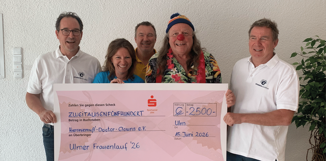 Five people standing indoors holding a large ceremonial check with text indicating a donation to a charity event. Beschreibung von KI generiert